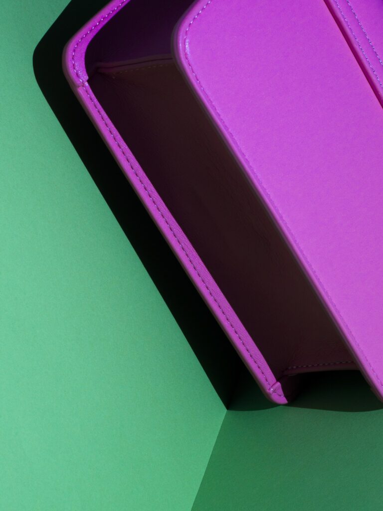 Minimalistische productfoto van een paarse handtas tegen een groene achtergrond. De strakke compositie en het spel van licht en schaduw benadrukken de geometrische vorm en de luxe afwerking van de tas. Een modern voorbeeld van productfotografie, vastgelegd door Jasper Mulder.