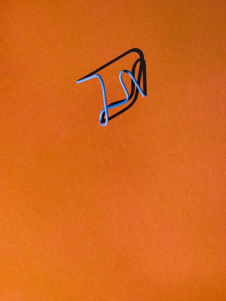 Minimalistische compositie van een vervormde blauwe paperclip op een warme oranje achtergrond. De schaduw voegt diepte en speelsheid toe, waardoor een grafisch en abstract beeld ontstaat. Vastgelegd in Amsterdam door Jasper Mulder.