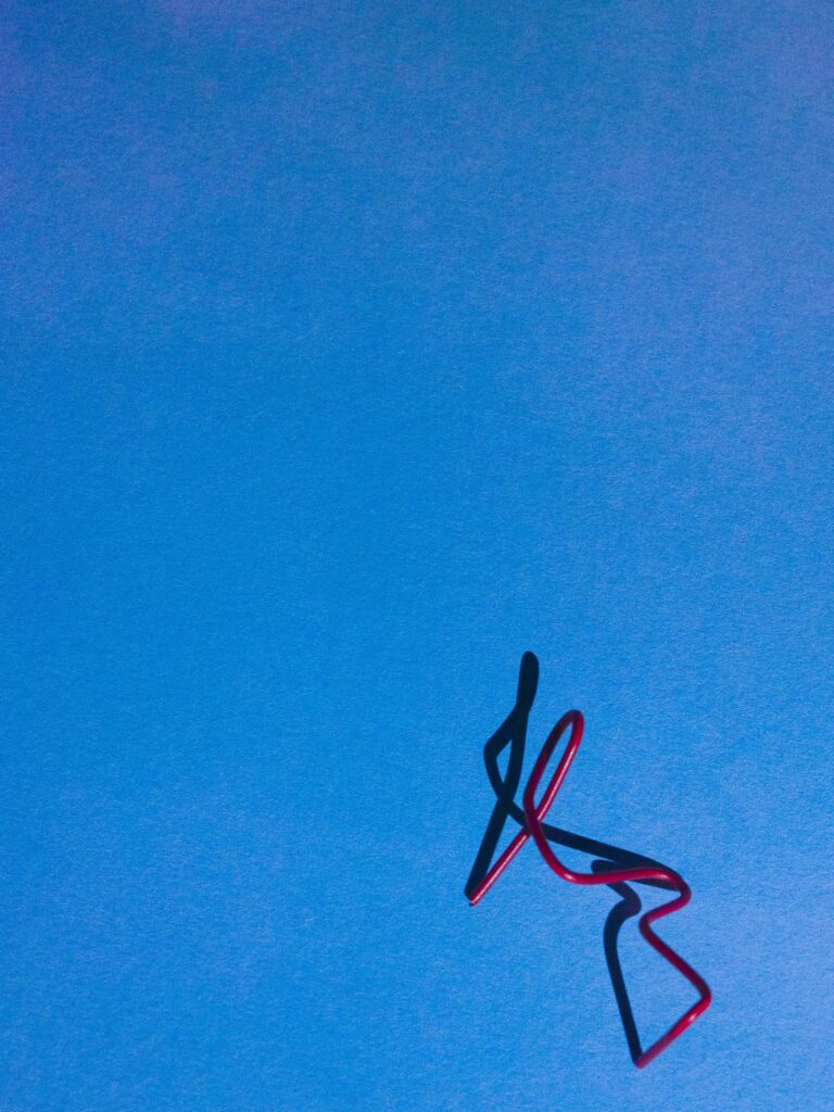 Minimalistische compositie van een vervormde rode paperclip op een felblauwe achtergrond. De speelse schaduw voegt dynamiek toe en benadrukt de eenvoudige, grafische esthetiek van het beeld. Vastgelegd in Amsterdam door Jasper Mulder.