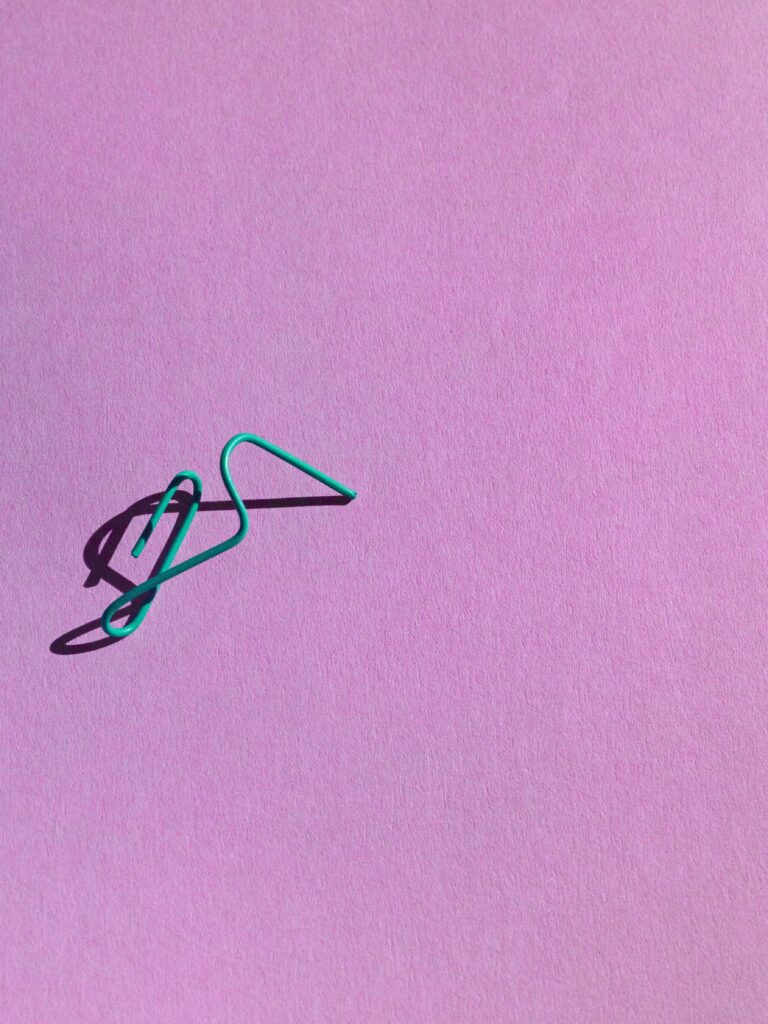Minimalistische compositie van een vervormde groene paperclip op een zachtroze achtergrond. De scherpe schaduw voegt diepte toe en benadrukt de eenvoud en speelsheid van het ontwerp. Vastgelegd in Amsterdam door Jasper Mulder.