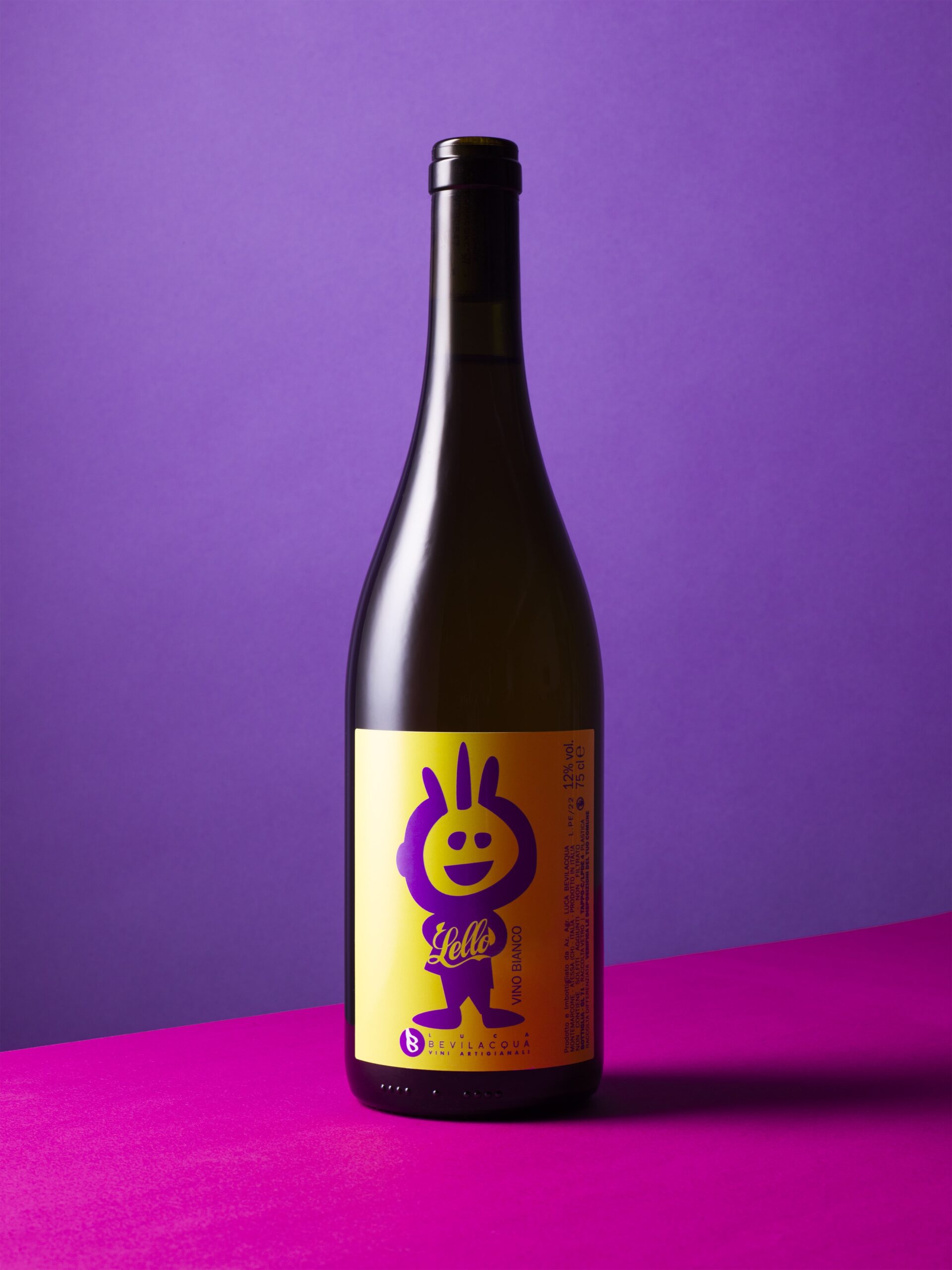 Productfoto van een fles witte wijn met een speels, kleurrijk etiket waarop een paars cartoonfiguurtje staat afgebeeld op een gele achtergrond. De fles staat rechtop op een felroze oppervlak met een paarse achtergrond, wat zorgt voor een opvallend en grafisch geheel. De fles draagt de naam 'Lello Vino Bianco' van het wijnhuis Luca Bevilacqua. Stijlvolle en kleurrijke productfotografie. Een voorbeeld van productfotografie, vastgelegd in Amsterdam door Jasper Mulder.