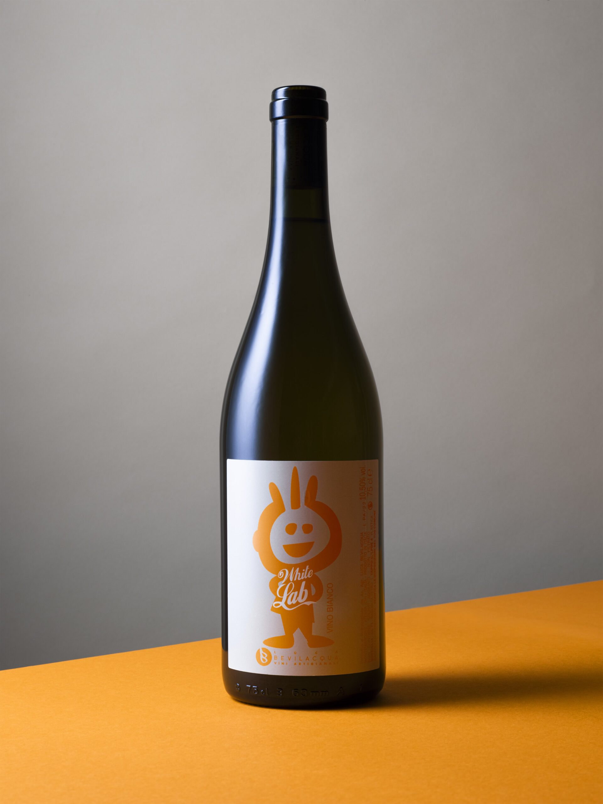 Productfoto van een fles witte wijn met een minimalistisch wit etiket waarop een oranje cartoonfiguur en de tekst 'White Lab' zijn gedrukt. De fles staat rechtop op een oranje oppervlak met een grijs-beige achtergrond. De wijn is van het merk Luca Bevilacqua. De strakke compositie en contrasterende kleuren benadrukken het speelse en moderne karakter van de productfotografie. Vastgelegd in Amsterdam door Jasper Mulder.