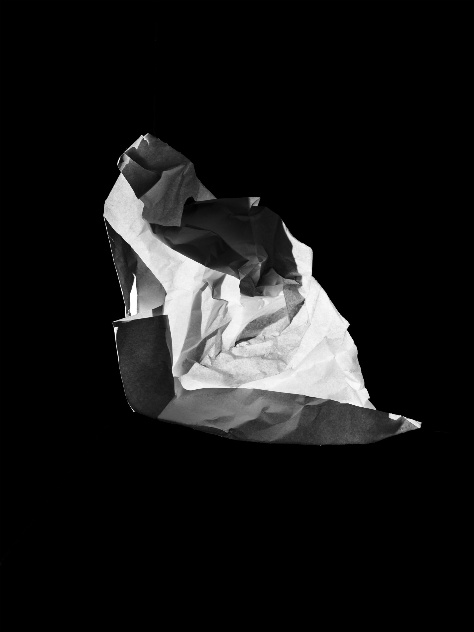Minimalistische en abstracte compositie van een zwevende, gekreukte papieren vorm op een diepe zwarte achtergrond. Het scherpe lichtspel benadrukt textuur en vorm, en creëert een sculpturale aanwezigheid. Dit werk vangt de essentie van vergankelijkheid en materialiteit, en vormt een abstraherende basis van het werk van Jasper Mulder. Vastgelegd in Amsterdam.