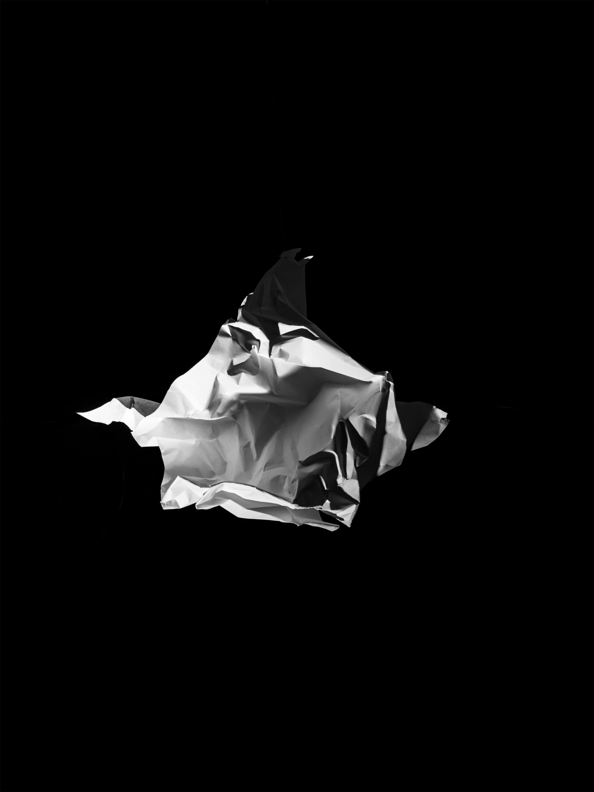 Minimalistische en abstracte compositie van een zwevende, gekreukte papieren vorm op een diepe zwarte achtergrond. Het scherpe lichtspel benadrukt textuur en vorm, en creëert een sculpturale aanwezigheid. Dit werk vangt de essentie van vergankelijkheid en materialiteit, en vormt een abstraherende basis van het werk van Jasper Mulder. Vastgelegd in Amsterdam.