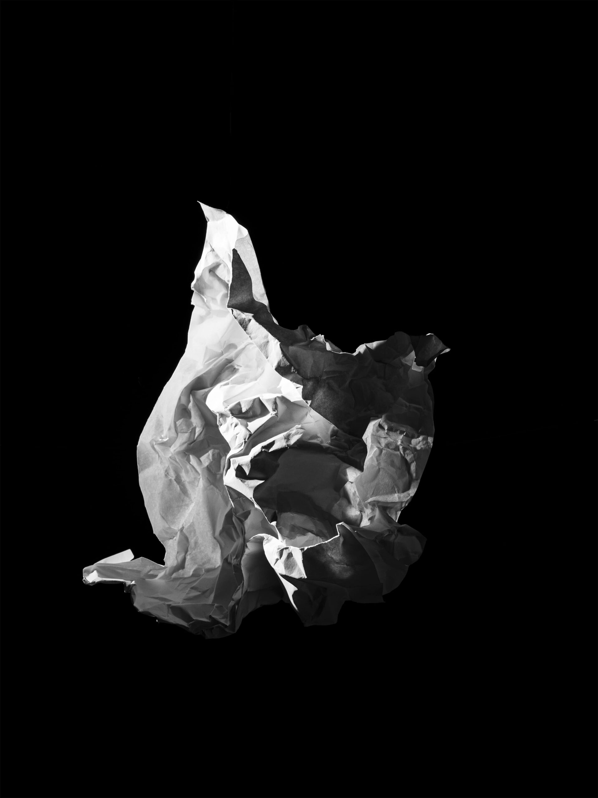 Minimalistische en abstracte compositie van een zwevende, gekreukte papieren vorm op een diepe zwarte achtergrond. Het scherpe lichtspel benadrukt textuur en vorm, en creëert een sculpturale aanwezigheid. Dit werk vangt de essentie van vergankelijkheid en materialiteit, en vormt een abstraherende basis van het werk van Jasper Mulder. Vastgelegd in Amsterdam.