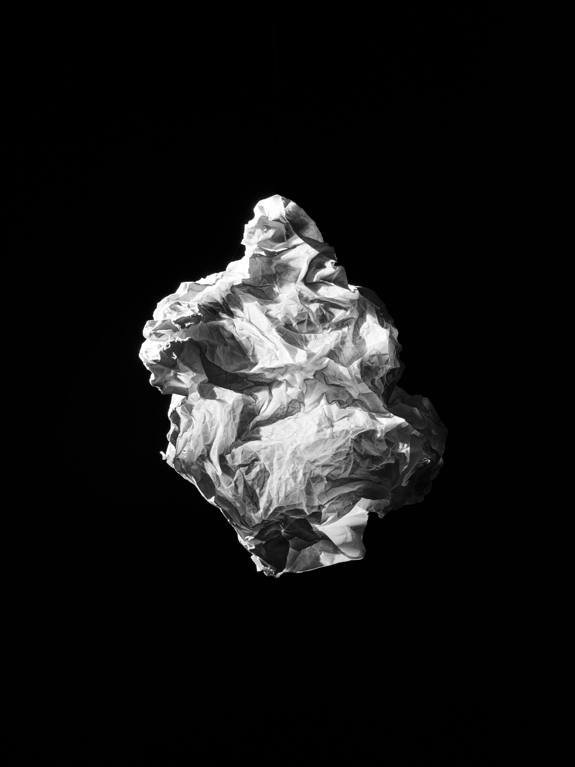 Minimalistische en abstracte compositie van een zwevende, gekreukte papieren vorm op een diepe zwarte achtergrond. Het scherpe lichtspel benadrukt textuur en vorm, en creëert een sculpturale aanwezigheid. Dit werk vangt de essentie van vergankelijkheid en materialiteit, en vormt een abstraherende basis van het werk van Jasper Mulder. Vastgelegd in Amsterdam.