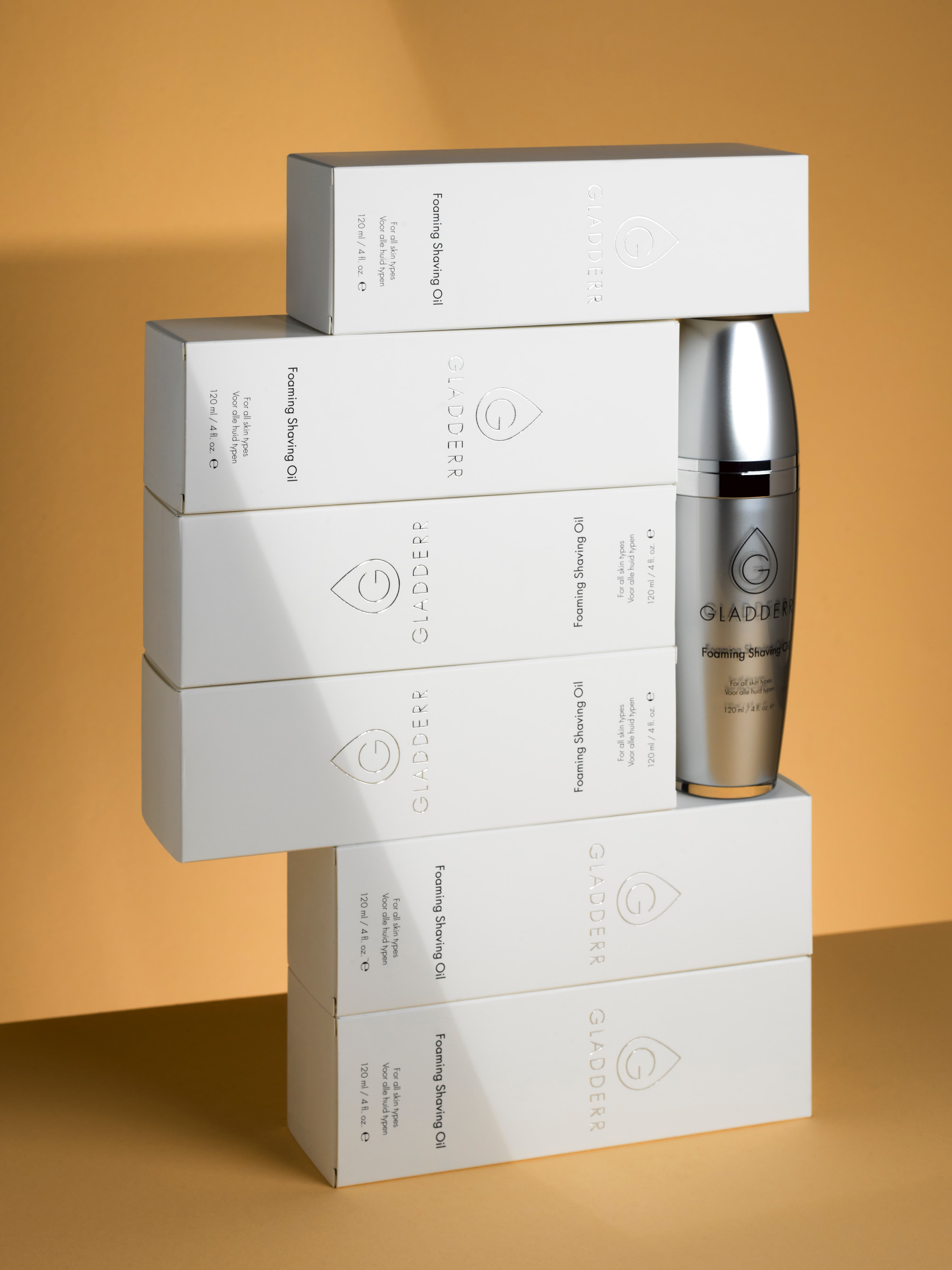 Productfoto van een Artistieke compositie van meerdere witte dozen Gladderr Foaming Shaving Oil, met één metallic fles centraal geplaatst tegen een zachte tan achtergrond. gefotografeerd in de studio.