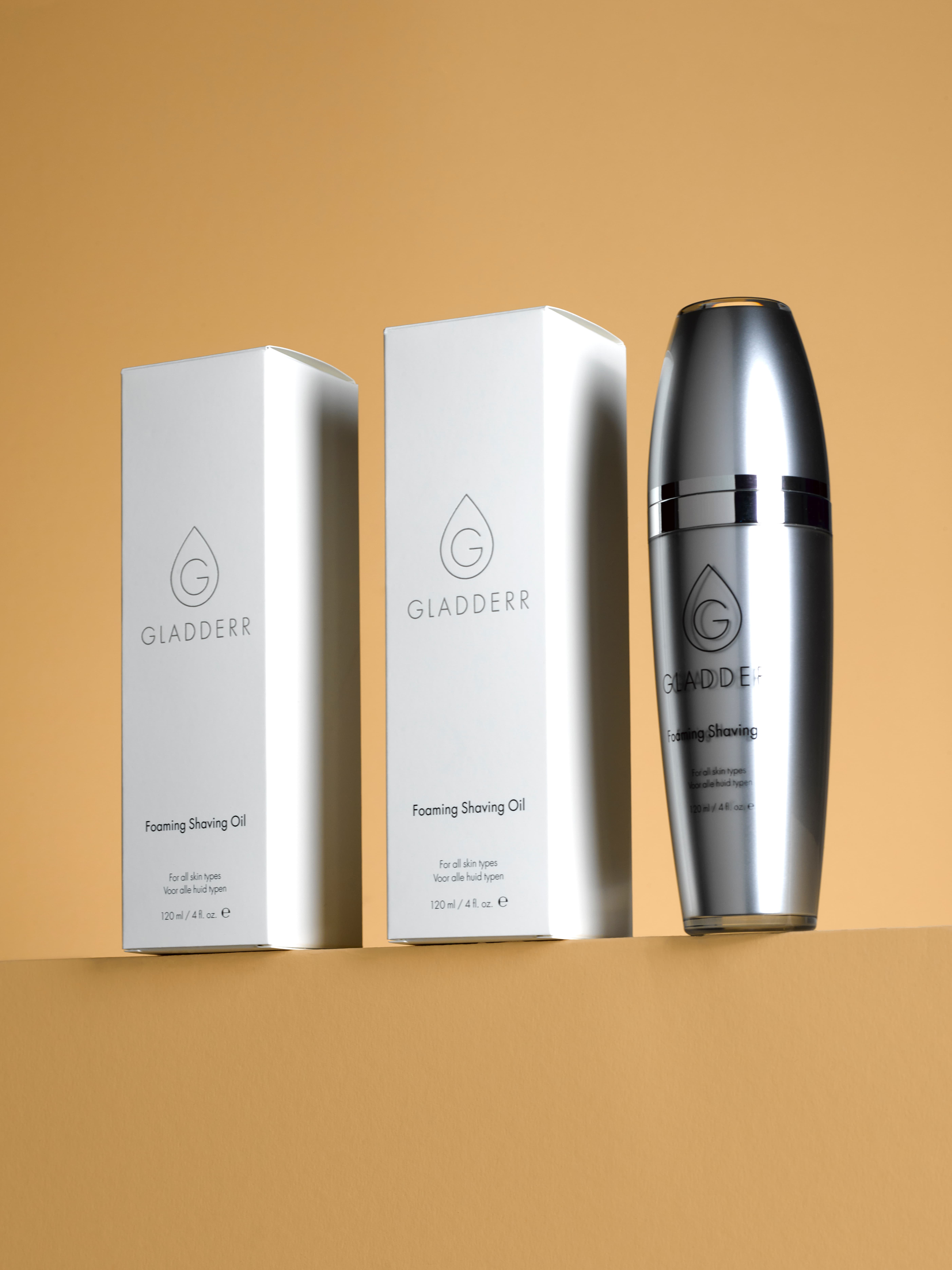 Productfoto van een luxe rvs fles en doos van Gladderr Foaming Shaving Oil, minimalistisch gepositioneerd op een vlakke ondergrond met warme tan tinten. Gefotografeerd in de studio.