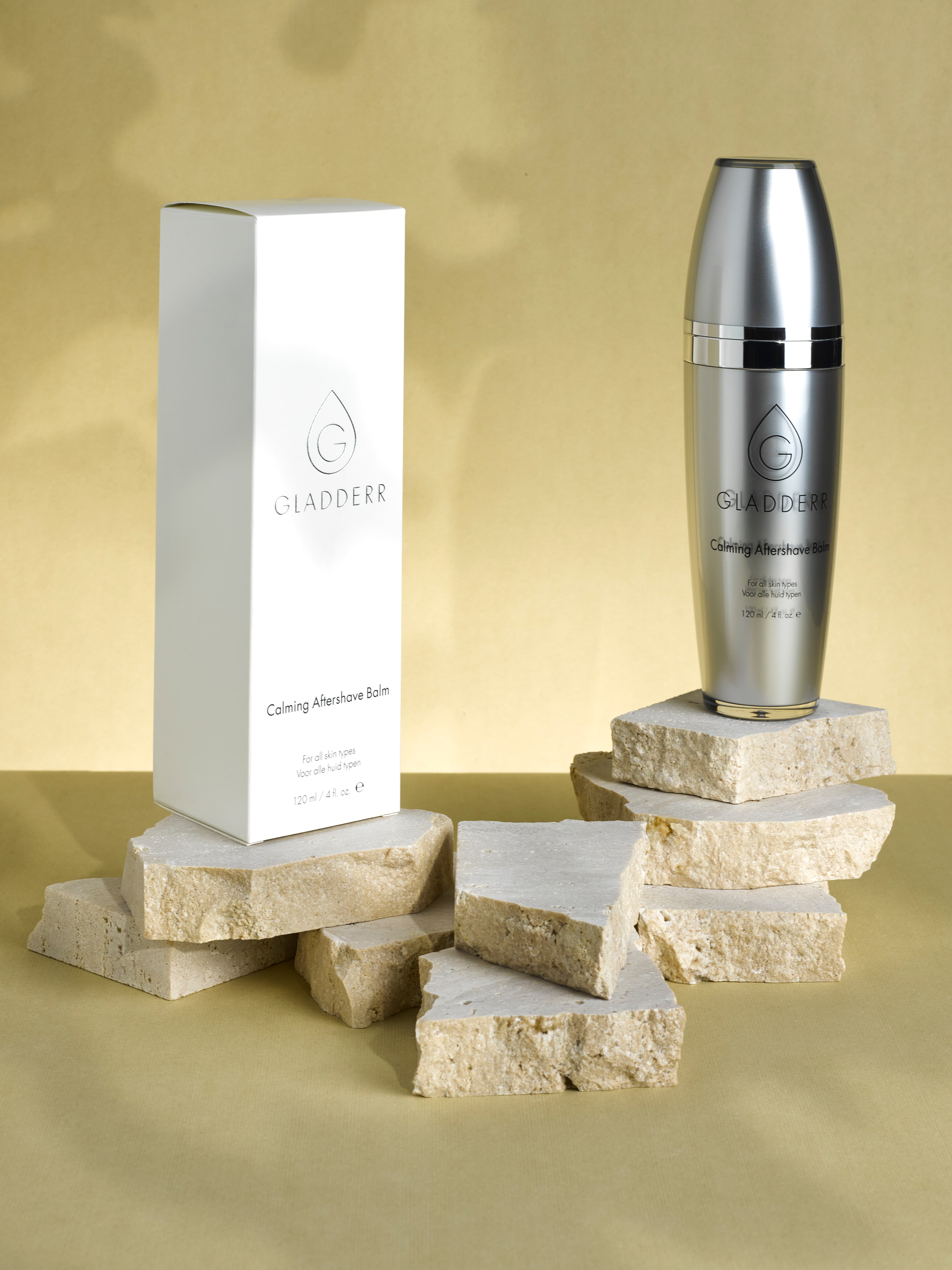 Productfoto van een luxe rvs fles en witte doos van Gladderr Calming Aftershave Balm, stijlvol gepresenteerd op gestapelde stenen met een neutrale beige achtergrond. Gefotografeerd in de studio.