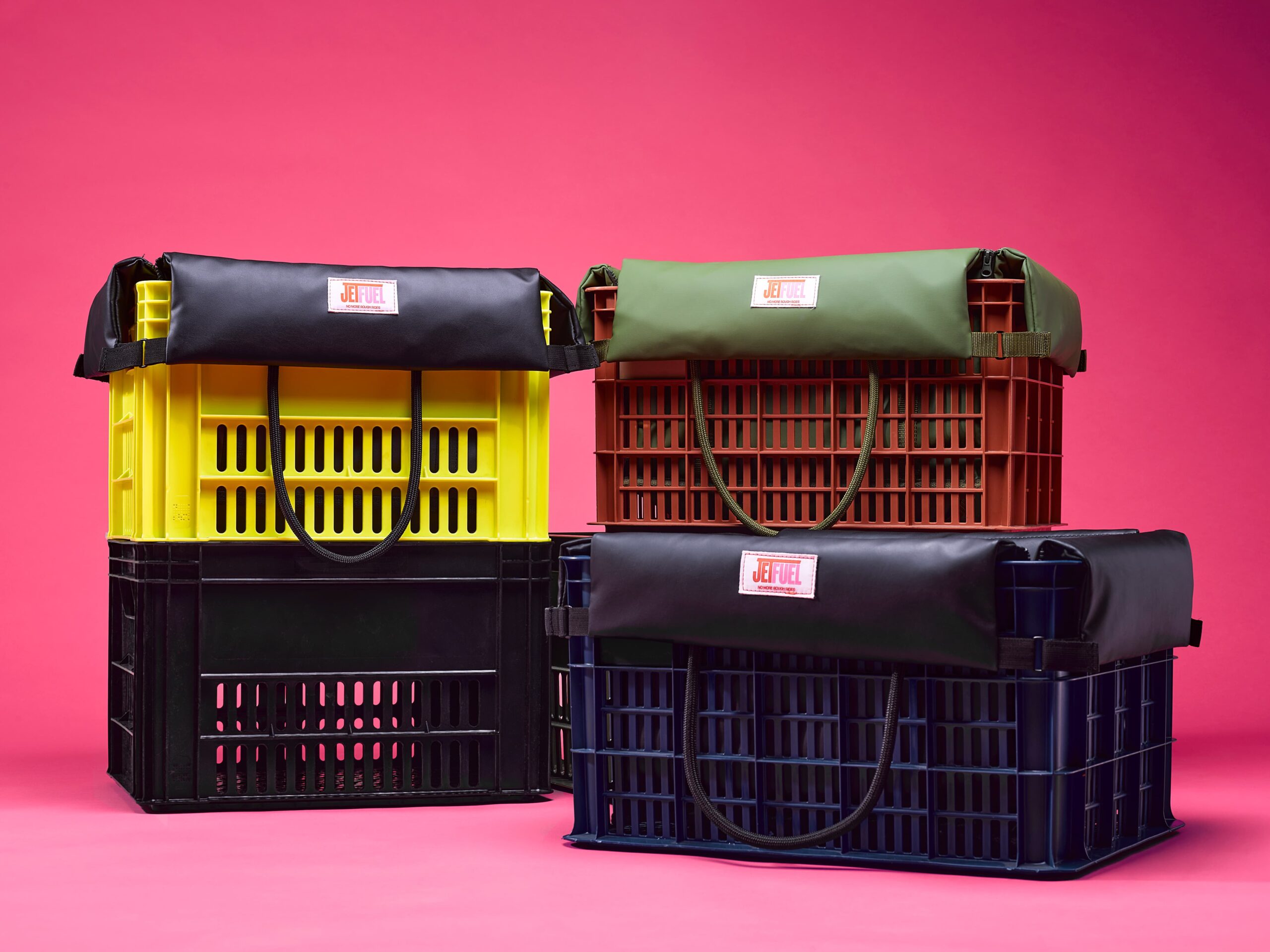 Vier JetFuel-krattenhoezen in zwart, olijfgroen en nogmaals zwart op gele, terracotta, zwarte en blauwe plastic kratten met touwhandvatten, tegen een roze achtergrond.