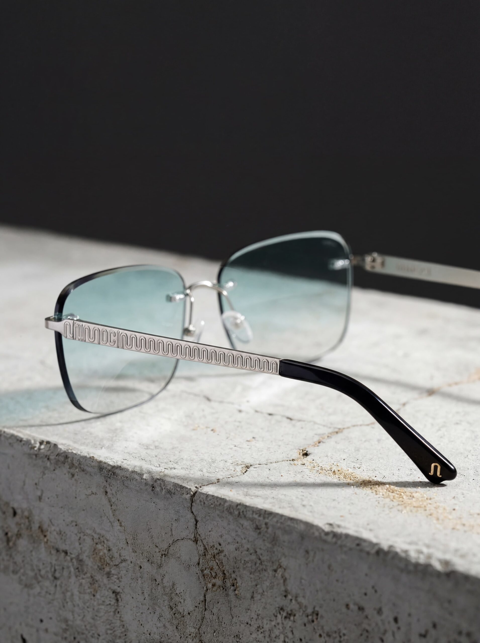 Moderne metalen bril met rechthoekige blauwgetinte lenzen en golvend zilveren pootjes, gestyled op een minimalistische betonnen ondergrond — premium productfotografie voor luxe eyewear‑merken.